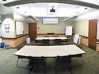 Three tables with 8 chairs each equidistant within the room, with a whiteboard and projector screen in the back of the room, bulletin board to the right wall, and a kitchenette to the left.