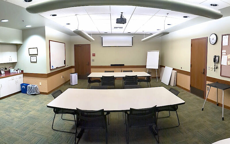 Three tables with 8 chairs each equidistant within the room, with a whiteboard and projector screen in the back of the room, bulletin board to the right wall, and a kitchenette to the left.