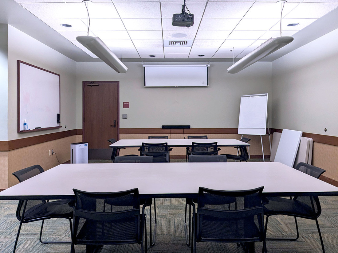 A close view of the far wall with the projection screen pulled down. Two tables with 6 chairs each are in front of the screen.