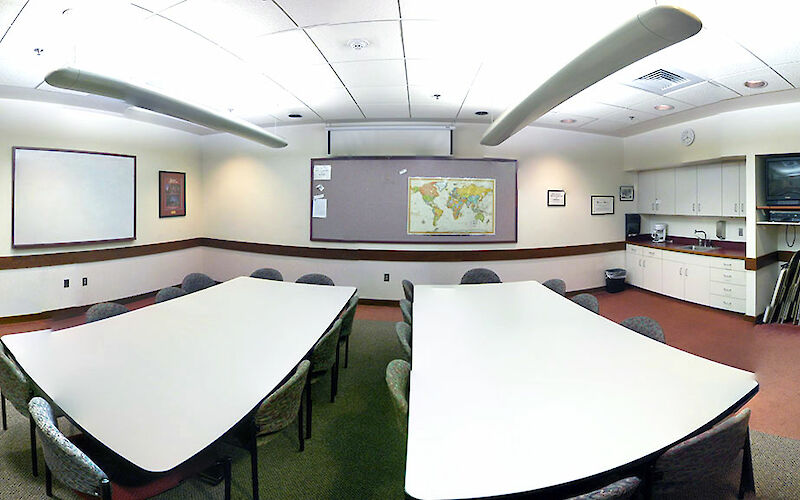 An overview of the entirety of the room: Two tables with 8 chairs each, a well-lit room with whiteboard and large bulletin board, and small kitchenette.