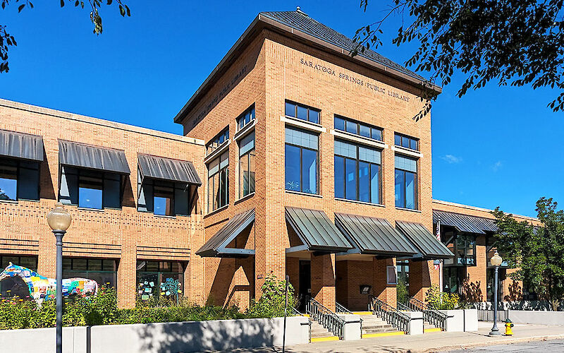 Shadows from overhead trees line the street in front of the main entrance to the Saratoga Springs Public Library on a day with a clear, blue sky.