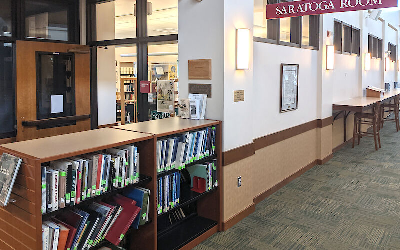 A door partially hidden by a waist high shelf of books with a sign to the right hanging from the ceiling to denote the entrance to the Saratoga (History) Room.