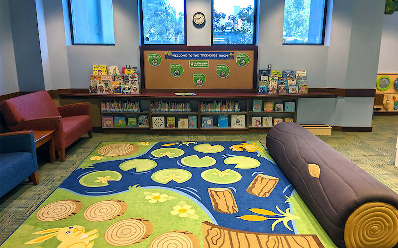 A play area with chairs, corkboard bulletin area, padded log seat, and decorative nature-themed carpet.