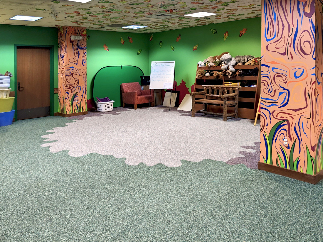 A diagonal view of the room from the entrance to the opposite corner. A wood bench, wooden shelving storage compartments, easel, and decorated columns are visible.