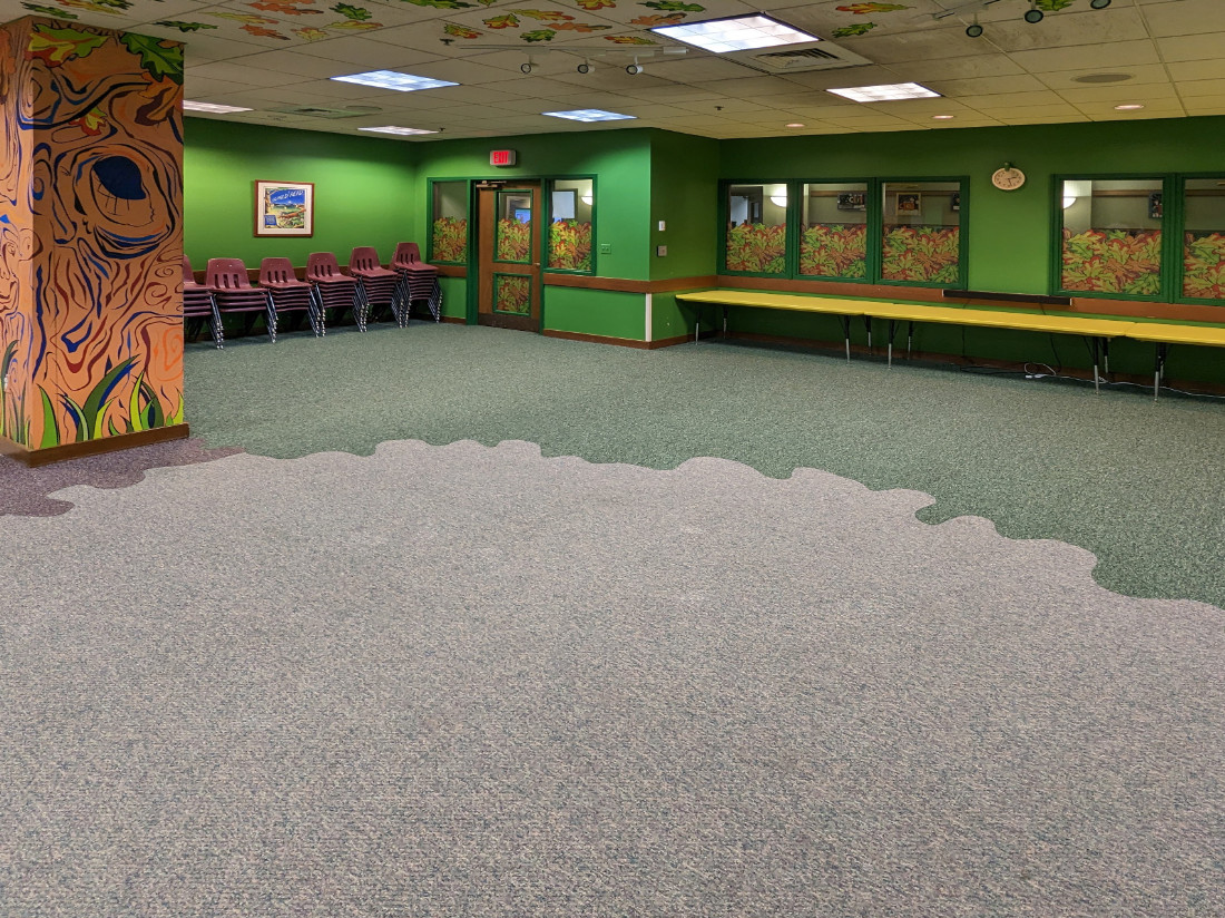 Viewing the room from the far corner to its entryway, diagonally. A column, long table, windows, and stacked chairs along a wall.