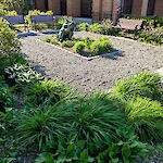 Thumbnail: A perspective view of a park area from the left-side of the entrance with benches, bushes, a center sculpture, and a crushed stone walkway.