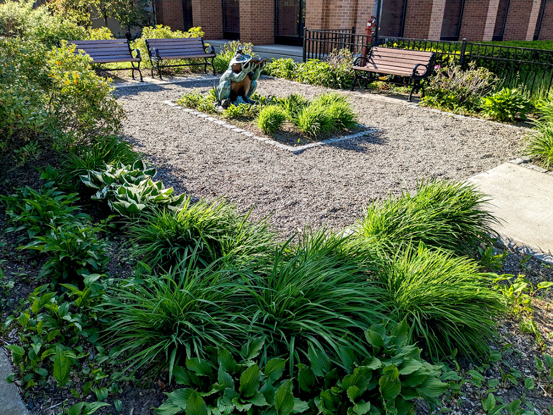 A perspective view of a park area from the left-side of the entrance with benches, bushes, a center sculpture, and a crushed stone walkway.