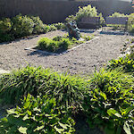 Thumbnail: A perspective view of a park area from the right-side of the entrance with benches, bushes, a center sculpture, and a crushed stone walkway.