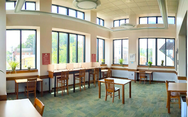 Tables with chairs around a room’s perimeter, against the walls. One lone table and chair in the center of the floor. Tall windows cover the majority of the walls.