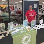 Thumbnail: A library staff member behind table with black tablecloth and green library logo with information on table