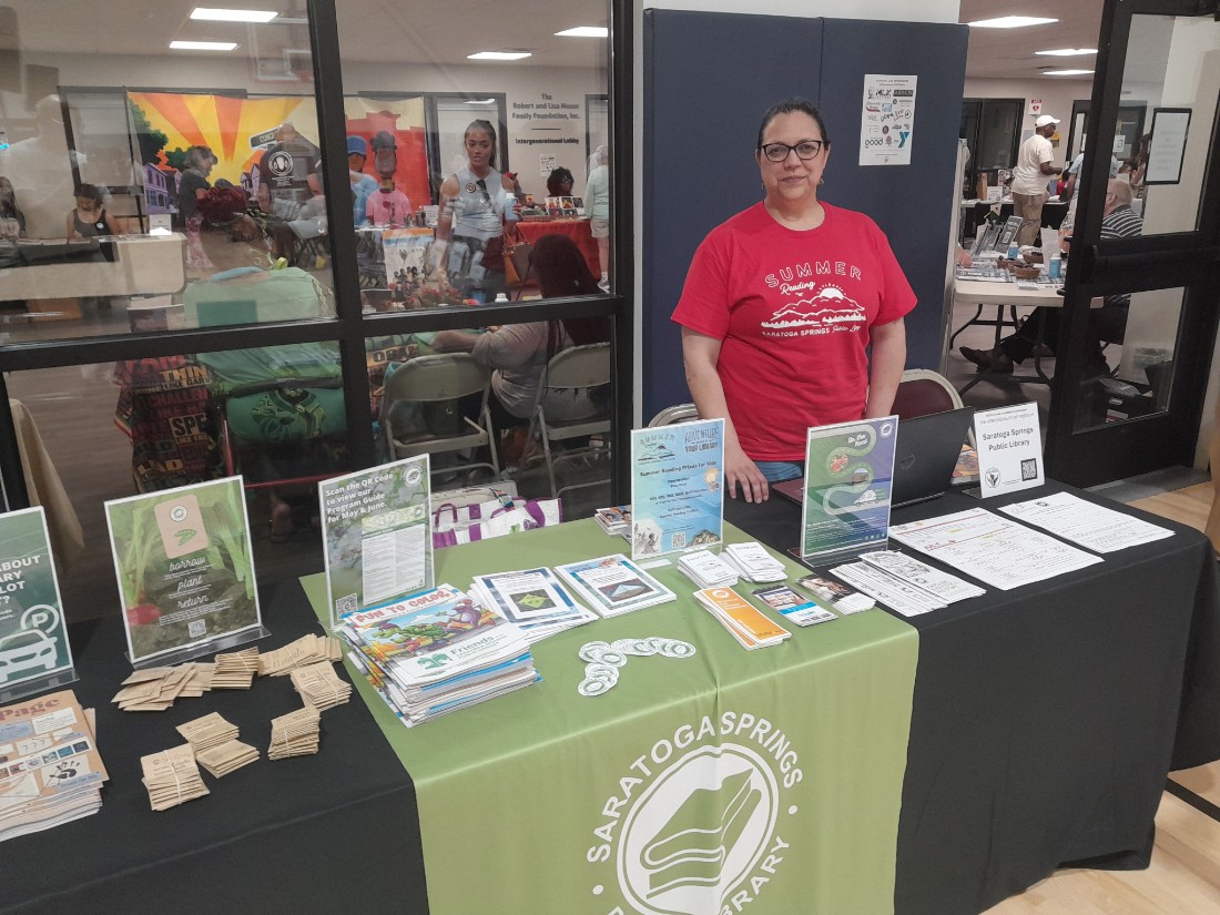 A library staff member behind table with black tablecloth and green library logo with information on table