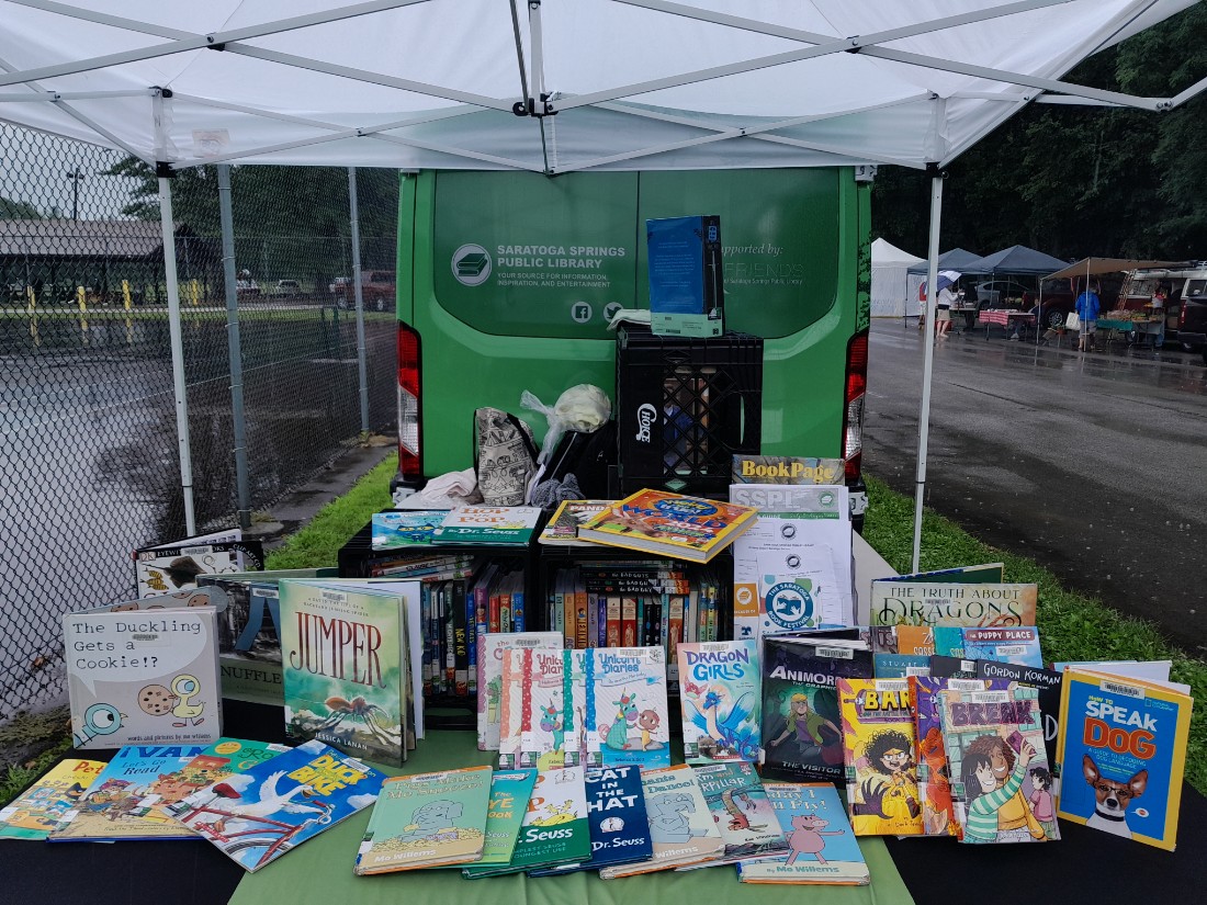 A panel van, decorated with the library’s colors of green and white.  Picture books display on a table with a black tablecloth and green runner.  White tent overhead.  Rain in the background.