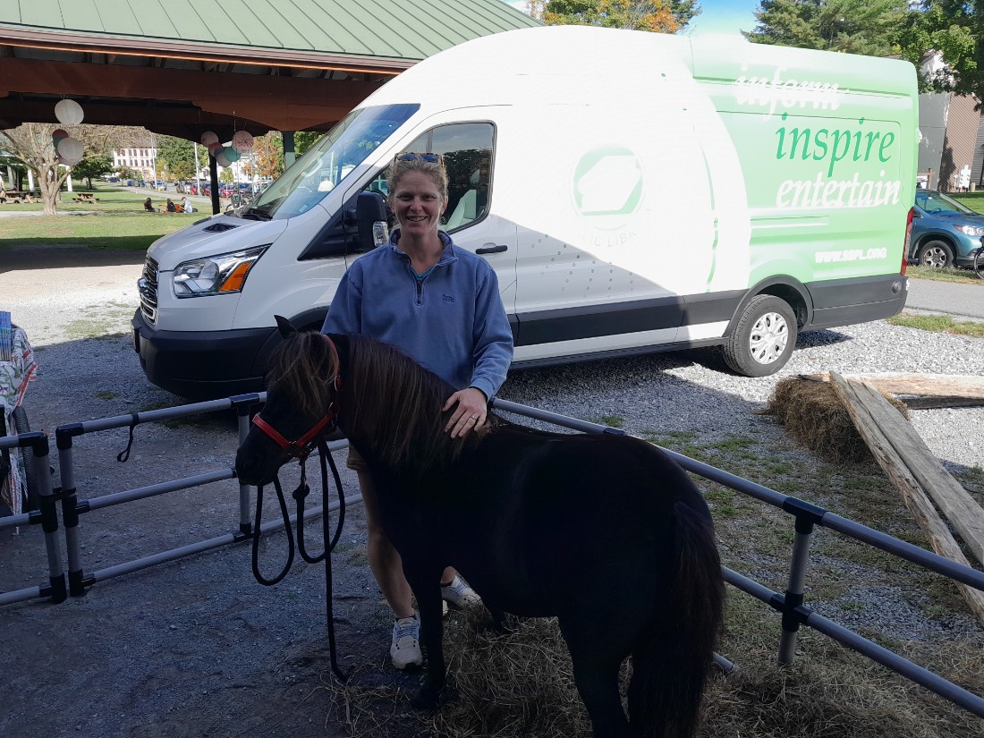 A woman in a blue shirt standing outside with a brown miniature horse in front of a panel van with a library logo