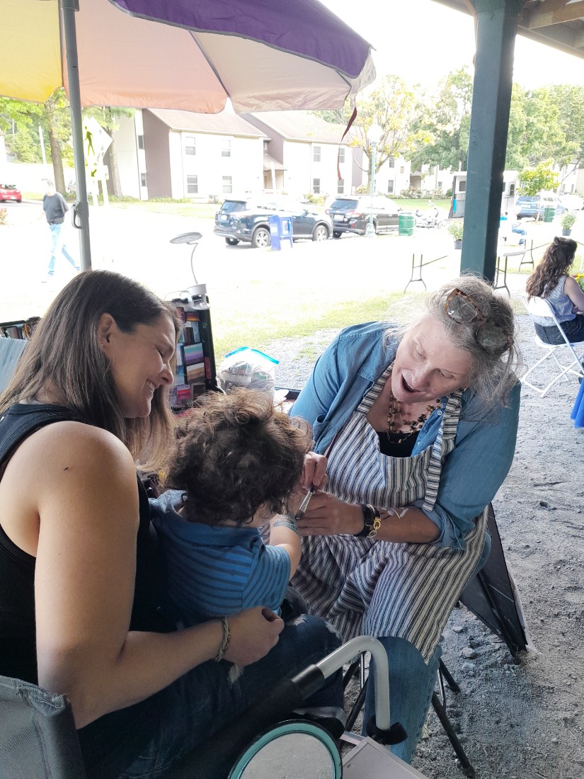 woman in chair with a happy face holding a child, another woman with apron and glasses painting child’s face