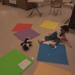 Thumbnail: color foam mats on floor with child coloring on mats.  cat stuffed animal next to child. sandwich board behind child with storyime time