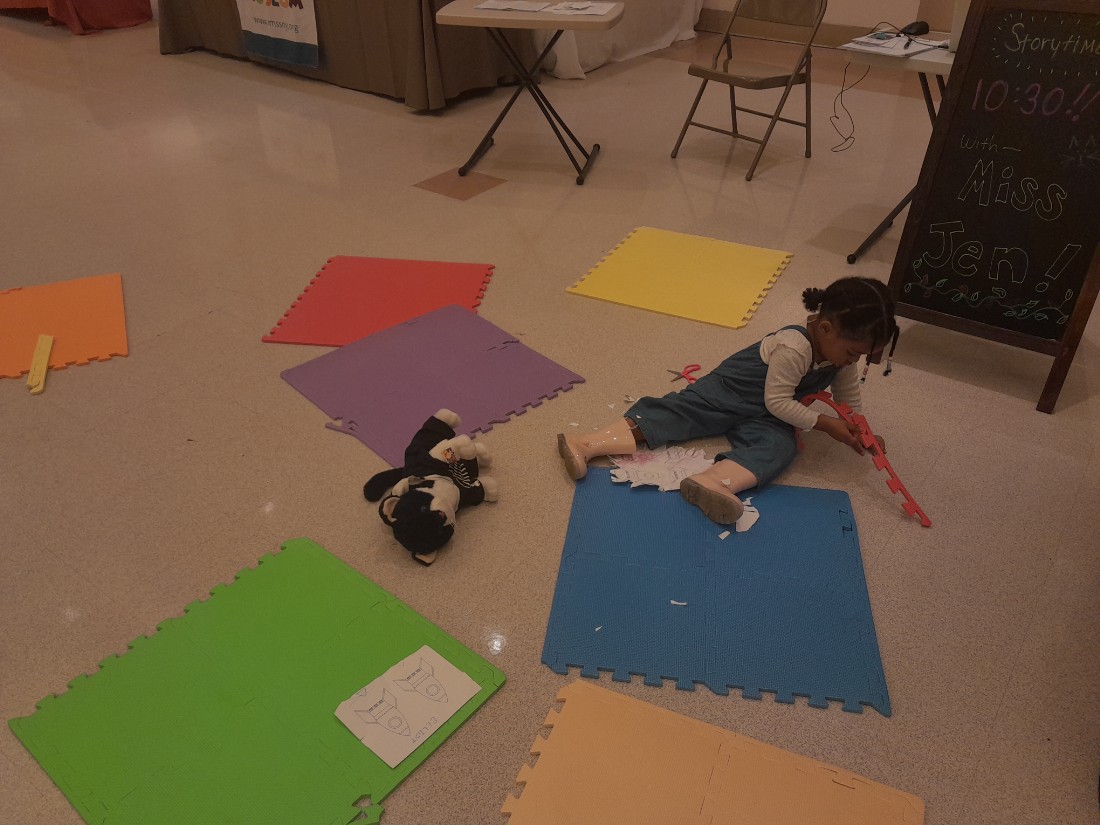 color foam mats on floor with child coloring on mats.  cat stuffed animal next to child. sandwich board behind child with storyime time