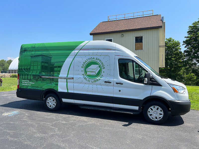 A panel van, decorated with the library’s colors of green and white showcasing a two-tone photo depicting the library all in green on the back half and the library logo in green on white on the front.