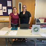 Thumbnail: Two people, one from the library, behind a white table with green library logo sign with information on table