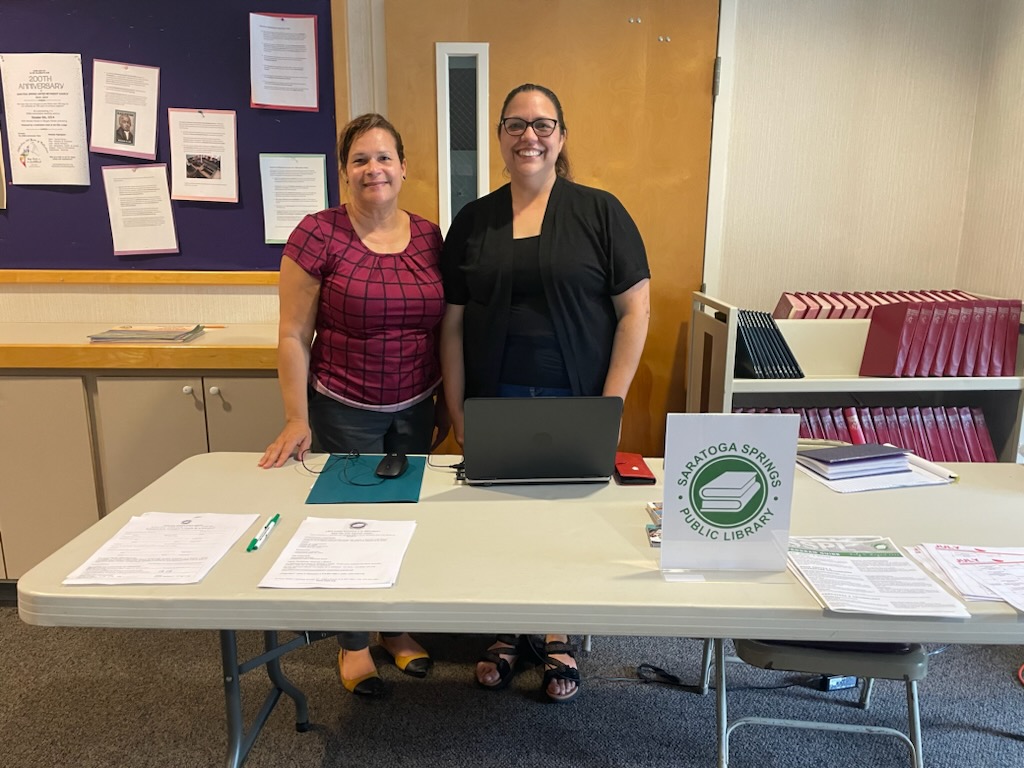 Two people, one from the library, behind a white table with green library logo sign with information on table