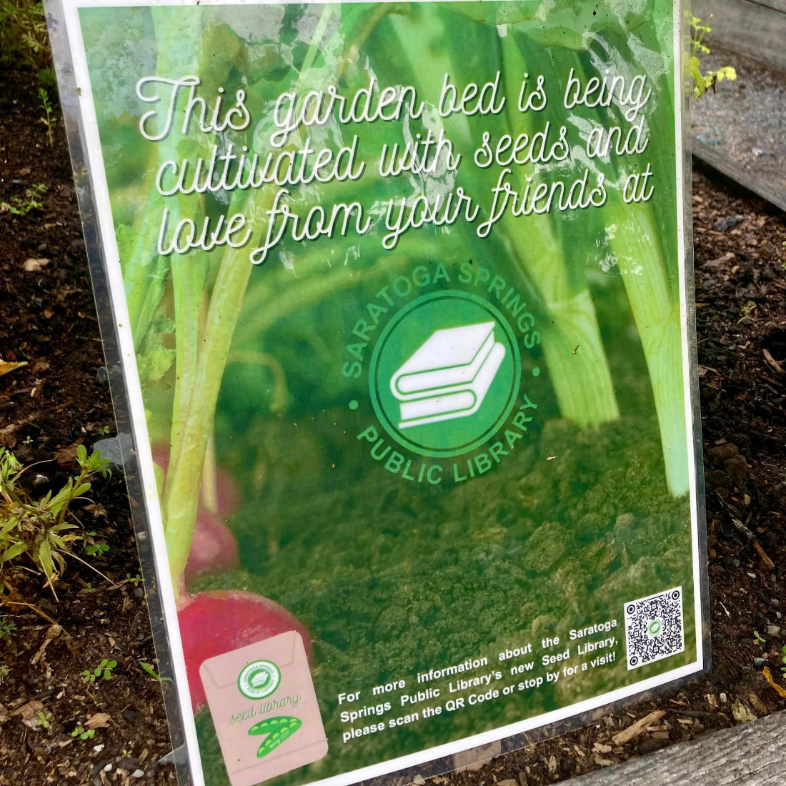 Text states This garden bed is being cultivated with seeds and lover from your friends at Saratoga Springs Public Library. Image shows garden plants growing a seed packet and the library logo